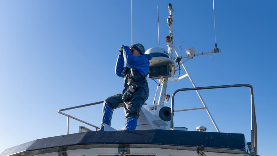 A surveyor in foul-weather gear standing on a boat's wheelhouse, scanning the water through binoculars.