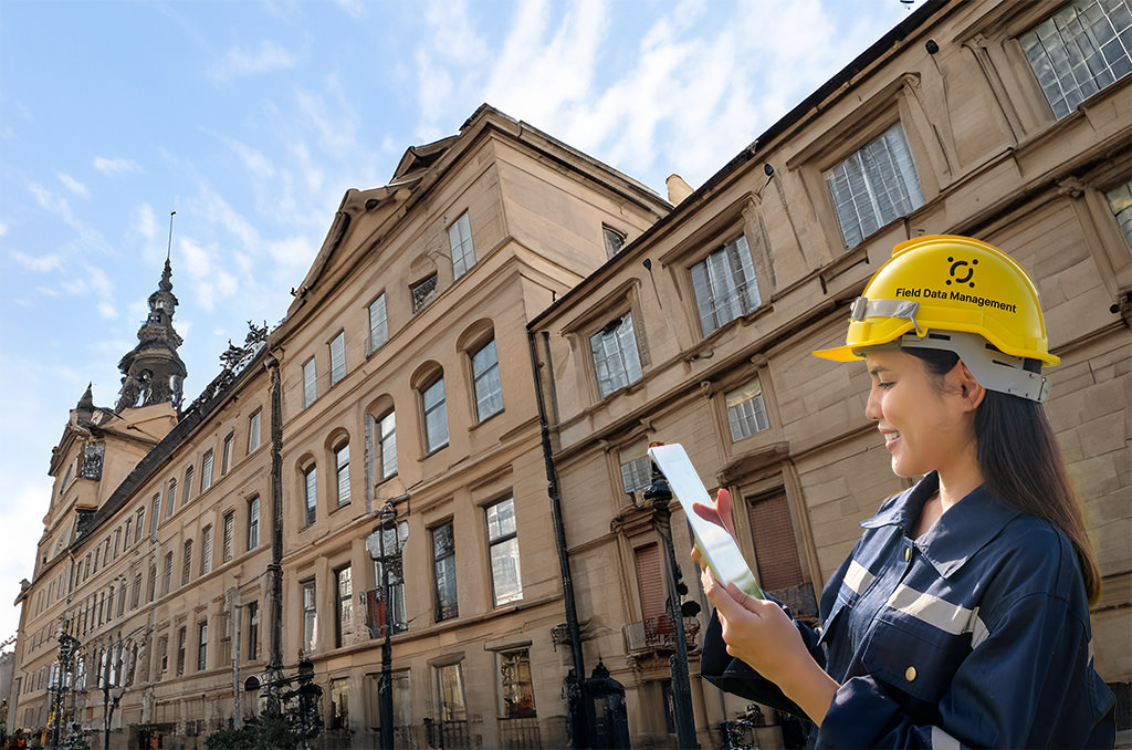 A field data management professional in a hard hat using a digital tablet in front of a classical architecture building for historic preservation documentation.