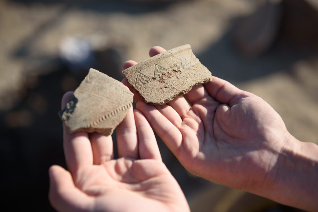 A pair of hands holding two pieces of pottery or ceramic shards, possibly artifacts, against a blurred outdoor background, representing archaeological fieldwork and artifact examination.