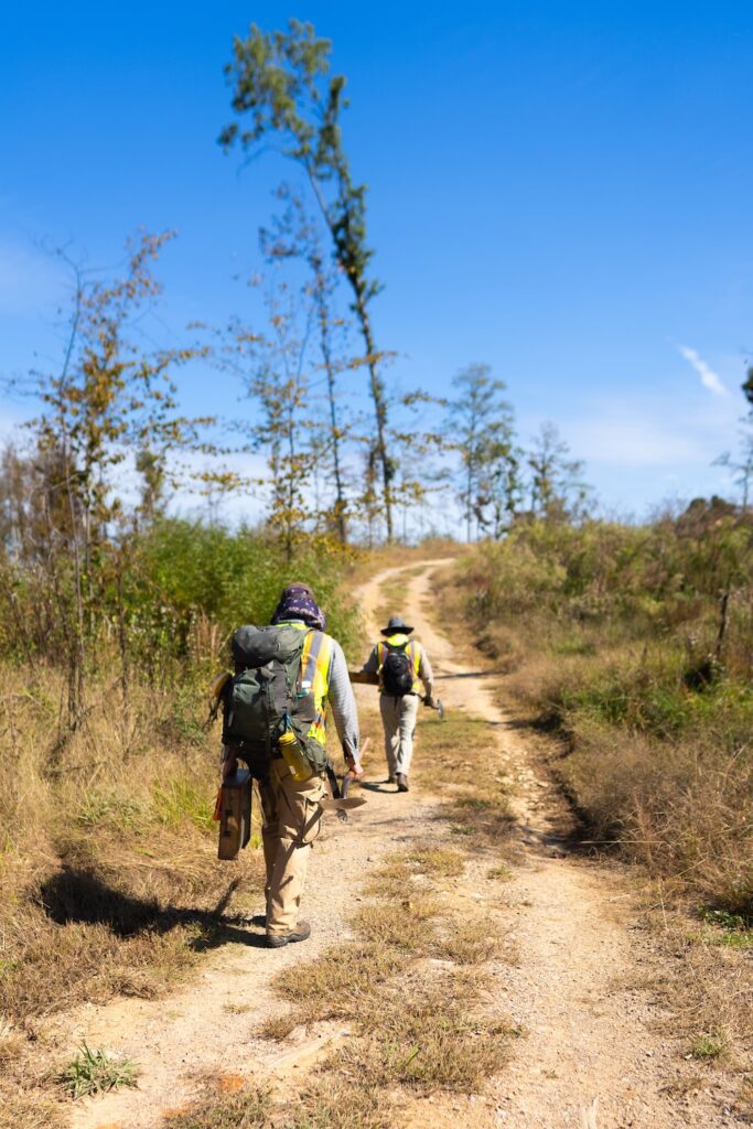 Two field technicians walk down a dirt road loaded with gear for CRM.