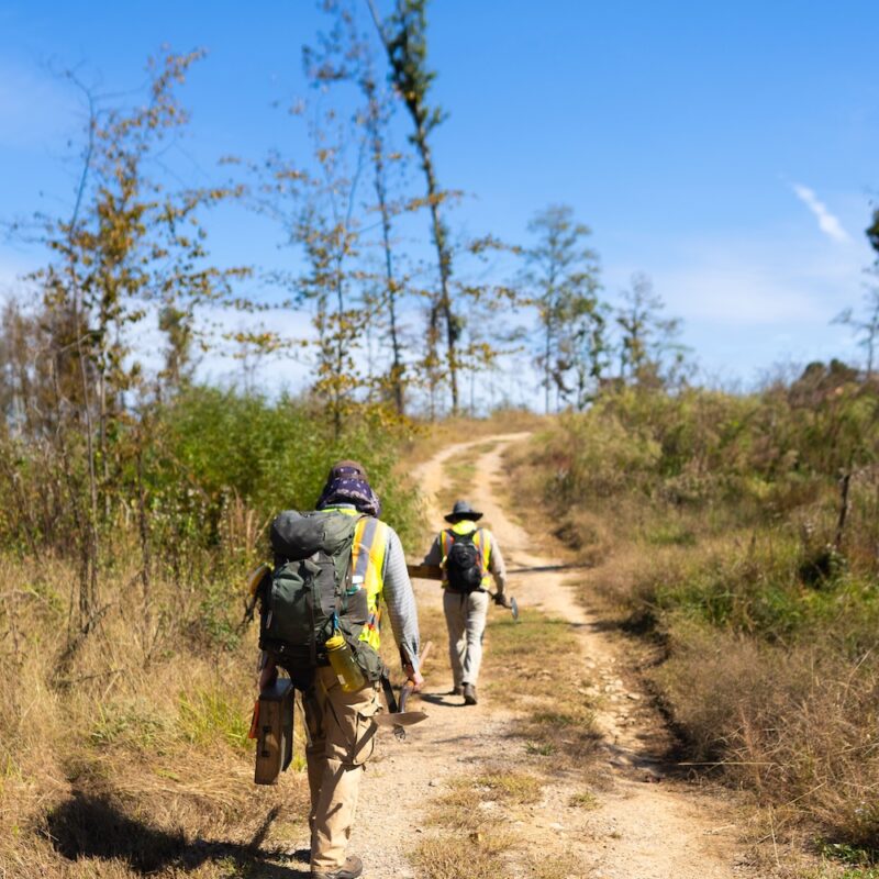 Two field technicians walk down a dirt road loaded with gear for CRM.